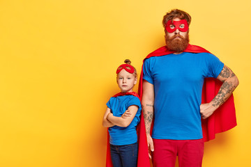 Photo of serious bearded man and self confident little child stands near with crossed arms, wears superhero costumes, show courage, put their life to risk, posses brave deeds and noble qualities © WHstudio Leushin N