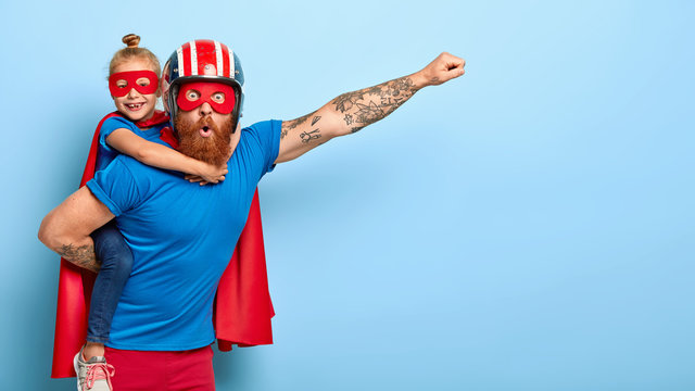 Horizontal shot of powerful man makes fly gesture, gives piggyback to small daughter dressed in superhero suit, have supernatural abilities, enjoy free time together. Real heroes defend you.