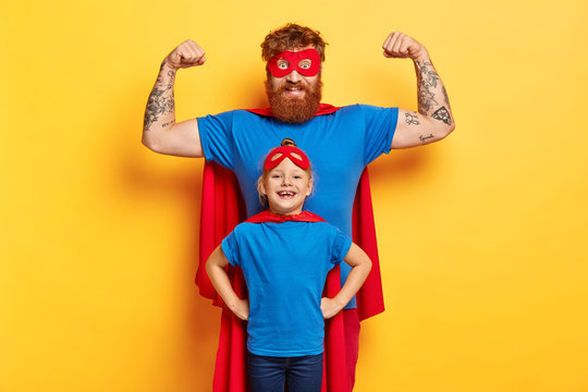 Family fun concept. Joyful strong father raises arms and shows biceps, ready to defend his daughter, stands back, imagine they are superheroes, wears costumes, isolated on yellow background.