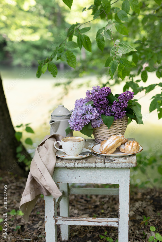 Wall mural Breakfast in the garden: eclairs, cup of coffee, coffee pot, lilac flowers in a basket.