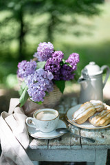 Breakfast in the garden: eclairs, cup of coffee, coffee pot, lilac flowers in a basket.