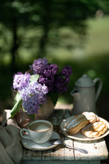 Breakfast in the garden: eclairs, cup of coffee, coffee pot, lilac flowers in a basket.