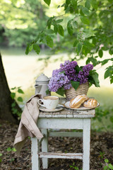 Breakfast in the garden: eclairs, cup of coffee, coffee pot, lilac flowers in a basket.