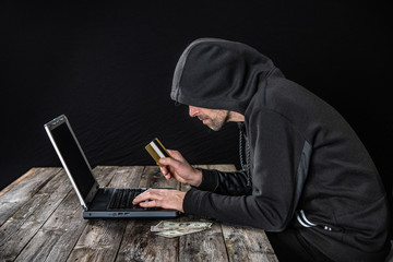 hacker in black hoody with laptop, bank card and dollar notes