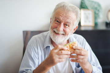 Seniors European man is sitting to eat a burger at home which looking at camera. Retired man is...