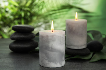 Fototapeta na wymiar Burning candles and spa stones on grey table against blurred green background