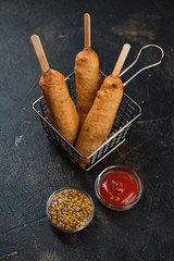 Deep-fried sausages covered in a dough and dipping sauces, vertical shot on a dark brown stone background, elevated view