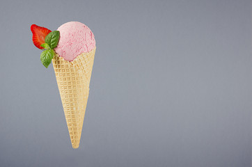 Delicious pink creamy ice cream scoop in crisp waffle cone with strawberry slices, mint leaf on grey background.