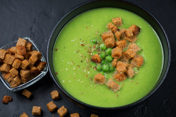 Bowl of cream-soup with green peas and croutons on a black stone background, close-up