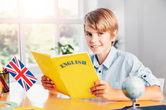 Young blonde smiling boy holding yellow english language book in light classroom