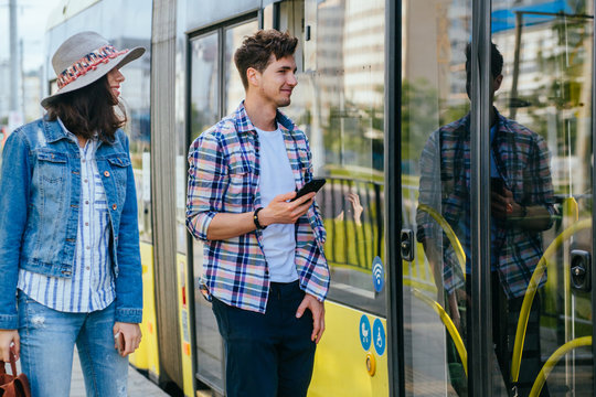 Lifestyle photo of young woman and man passengers holds phones waiting while opening door to tram. Portrait of female traveler with backpack talking with handsome guy on tram stop.
