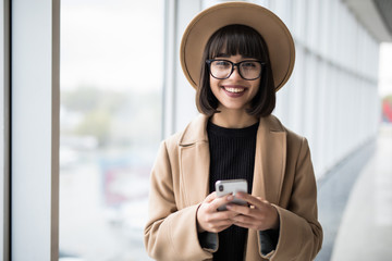 Young businesswoman wear in coat and hat with glasses talking on mobile phone while standing by window in office.
