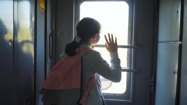 little teenage girl is a backpacker traveling by train. travel transportation railroad concept. tourist school girl in the train wagon with backpack looking out the window waving his hand lifestyle