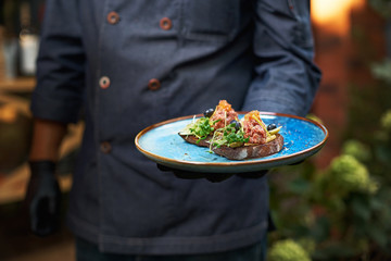 delicious raw beef tartare on bruschette, the dish is held in hand by a cook in a blue uniform.