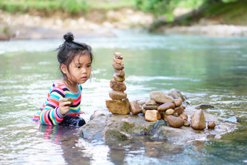 Asian child or kid girl learning sort or stack pebble stone tower or pyramid on water for balance training and meditation with nature relax on summer holiday travel at stream or river in forest spa