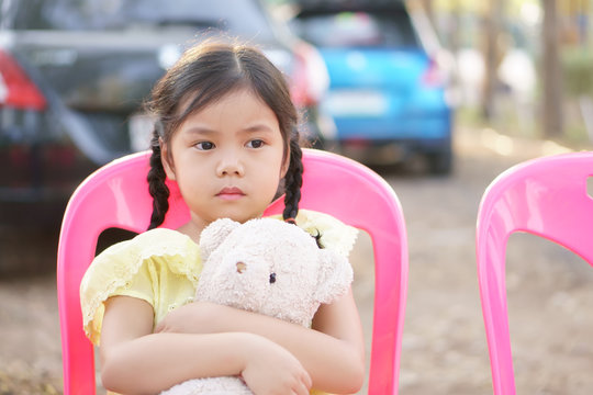 Asian child cute or kid girl pigtail sad with miss and lonely with hug teddy bear doll to lovely toy on street or city park with car and vacant sit on pink chair