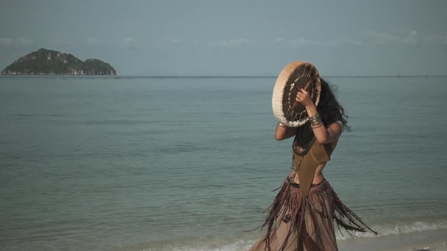 Attractive ethnic tribe girl dancing with a drum whirling around herself on the beach with sea and island background