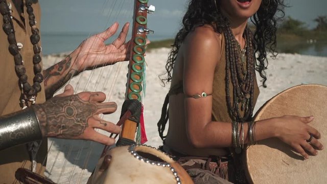 Close up of man playing on ethnic string instrument on sandy beach next to woman playing on drum
