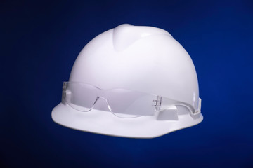 White generic protective construction helmet with a plastic transparent safety glasses on the cap against a blue studio background. Floating workers hat and spectacles.