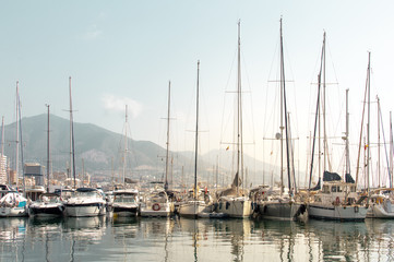 Fototapeta na wymiar Sailboats and yachts in the dock reflected in the water with the city and mountains in the background. Landscape of Costa del Sol (Fuengirola), Spain.