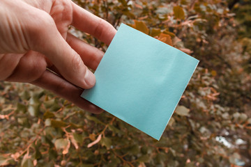 empty sheet of square blue paper in a male hand. fingers hold empty sticker, trees in a background, selective focus.