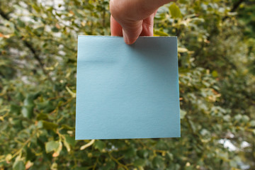 young man's fingers hold a square blue piece of sticky note, green trees in background. fingers hold empty blue square paper card