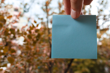 fingers hold empty blue square paper sticky note, trees in a background. square paper with space for text on a white.