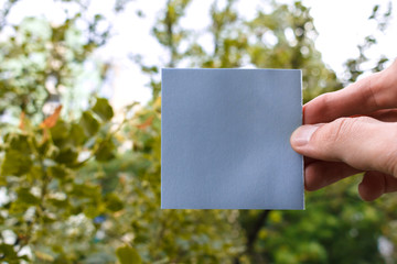 fingers hold empty blue square sticky note, green trees in a background. square paper with space...