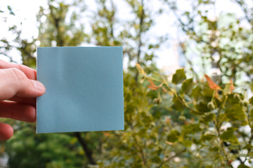 young man's fingers hold a square piece of blue paper, green trees in background