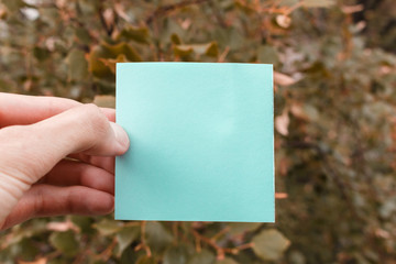male hand holding an empty blue sticker. square paper with space for text in the hand of a young man, selective focus. fingers hold empty blue square sticky note.