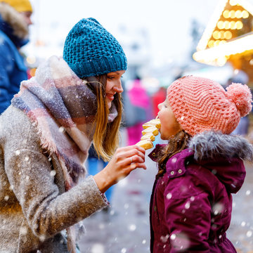 Young mother and daughter eating white chocolate covered fruits on skewer on traditional German Christmas market. Happy girl and woman on traditional family market in Germany during snowy day