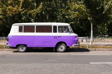 purple Retro van standing on the road near the fence, a park in the background. Classic wagon car is ready for summer vacations and trips to the sea