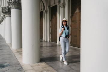 full length of young female traveler sightseeing in foreign city on weekend overseas. trendy woman tourist with rucksack and camera walking in corridor in rome style monument during summer adventure