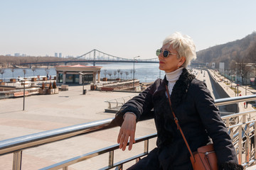 Fototapeta premium fashionable adult european woman in black glasses posing at the rail against the background of the highway and the bridge across the river. middle-aged woman tourist and traveler.