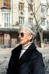 beautiful gray-haired lady in sunglasses stands on the street near the road, city background, profile portrait. fashionable middle-aged woman photographed in profile on the street, vertical image.