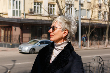 beautiful gray-haired lady in sunglasses stands on the street near the road, city background, profile portrait. fashionable middle-aged woman photographed in profile on the street.