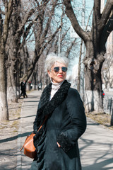 street fashion style portrait of stylish caucasian middle-aged woman with gray hair wearing sunglasses. gorgeous gray-haired caucasian woman with glasses standing on the street, vertical image.