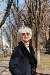 fashionable gray-haired caucasian woman in sunglasses and warm black coat posing for the camera on street boulevard. glamorous middle-aged european woman on a street, vertical image