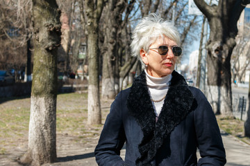 Fototapeta premium stylish caucasian woman with gray hair in a black coat and sunglasses standing on the alley in the city. fashion and style of middle-aged women.