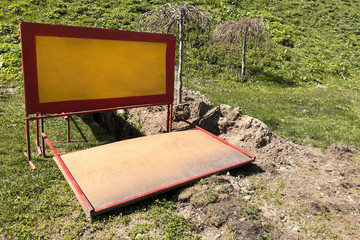 Orange-red wooden construction warning barricades fence the place where the trench is dug and pipe laying, green grass of the park in background, summer weather.