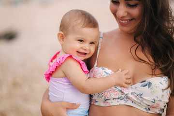 Portrait of Mother hold baby girl on hands on the beach in the sea. Mom and daughter in swimsuit. Close up of beautiful baby smile