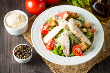 Fresh salad made of tomato, ruccola, chicken breast, eggs, arugula, crackers and spices. Caesar salad in a white, transparent bowl on wooden background