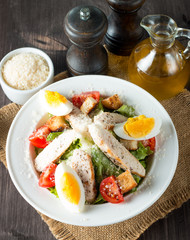 Fresh salad made of tomato, ruccola, chicken breast, eggs, arugula, crackers and spices. Caesar salad in a white, transparent bowl on wooden background