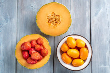 Top view of strawberry in melon and kumquat in a coconut bowl and a half of melon on white wooden background