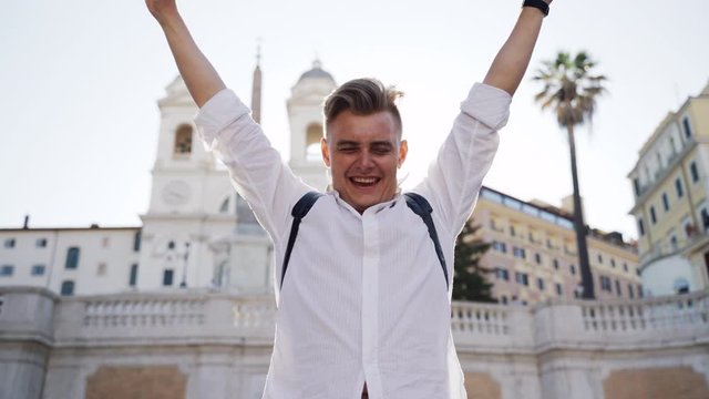 Confident young man lifting his hands up and showing the sign of victory and success. Happy tourist discovering world and enjoying vacation. Ancient city architecture and romatic atmosphere flying in