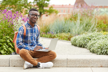 Fototapeta premium Full length portrait of African student smiling at camera while using laptop outdoors in college campus, copy space