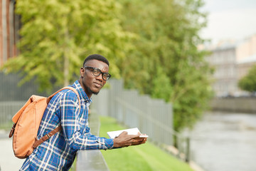 Waist up portrait of African student looking at camera while reading book outdoors in beautiful city, copy space