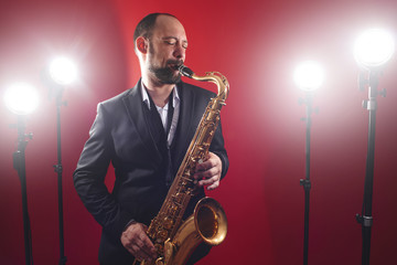 Obraz na płótnie Canvas Portrait of professional musician saxophonist man in suit plays jazz music on saxophone, red background in a photo studio