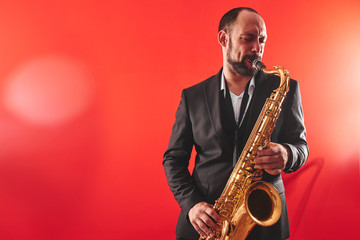 Fototapeta na wymiar Portrait of professional musician saxophonist man in suit plays jazz music on saxophone, red background in a photo studio