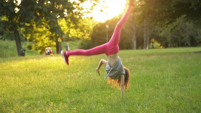 Happy Little Girl In Dress Is Jumping On Park Meadow And Making Acrobatic Wheel. Summer Outdoor Activities. Slow Motion.